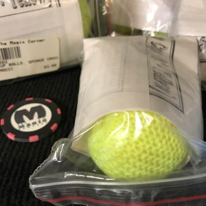 May include: A close-up of a yellow sponge ball in a clear plastic bag. The bag is sealed with a red zipper. The ball has a textured surface. The bag is labeled "The Magic Corner" and "Sponge Balls".