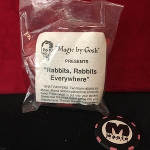 May include: A red plastic bag with a white label that reads "Magic by Gosh" presents "Rabbits, Rabbits Everywhere". The label describes a magic trick with two foam rabbits. A black and pink poker chip with the word "MAGIC" in white letters is in the foreground.