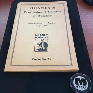 May include: A tan-colored paper catalog titled Heaney's Professional Catalog of Wonders, featuring text for Magical Goods, Illusions, and Stage Acts, with a logo of a magician and a rabbit. Beside it sits a round black and pink poker-style chip.