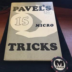 May include: A softcover book titled Pavel's 15 Micro Tricks with a beige cover featuring black text and a graphic of a hand holding a circle. Next to the book is a round black and pink poker-style chip with a white M and the text The Magic Company.