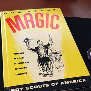 May include: A yellow book titled "Cub Scout Magic" with a cartoon illustration of a magician performing a magic trick. The book lists "Tricks, Magic, Puzzles, Stunts, Games" and is published by "Boy Scouts of America".