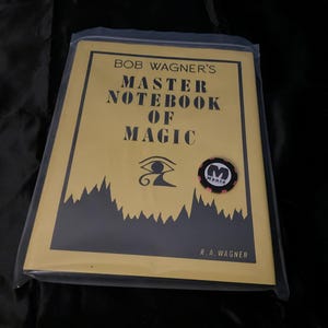 May include: A yellow book titled Bob Wagner's Master Notebook of Magic, enclosed in a clear plastic sleeve. The cover features black text, an Eye of Horus symbol, and a small circular black and white poker chip with the letter M and the word Magic.