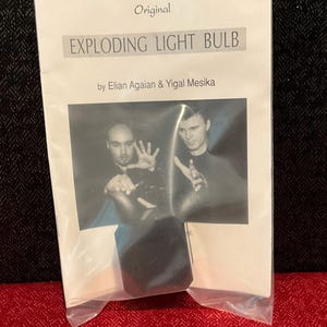 May include: A clear plastic package containing a black object and a printed card. The card reads "Original EXPLODING LIGHT BULB" with the names Elian Agaian & Yigal Mesika. A black and pink Magic chip is in the foreground.