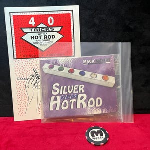 May include: A magic trick set including a white instruction booklet titled 40 Tricks with a Hot Rod by Scott K. Anderson, a Magic Makers Silver Gem Hot Rod in a clear plastic bag, and a black and pink circular token with a white M logo.