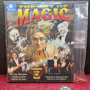 May include: A book titled The Art of Magic, the companion to the PBS special, featuring a collage of magicians and performers on the cover. The book is enclosed in a clear plastic sleeve and is accompanied by a small round black and white token.