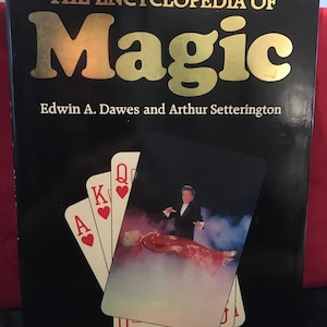 May include: A black hardcover book titled The Encyclopedia of Magic by Edwin A. Dawes and Arthur Setterington. The cover features gold text and an illustration of playing cards, including an ace, king, and queen, with a magician performing.