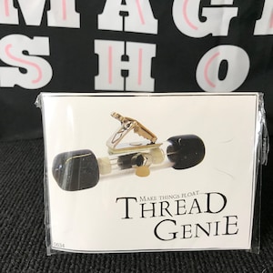 May include: A Thread Genie magic prop in clear plastic packaging. The device features a clear tube with black end caps, a metal clip, and internal thread mechanism. The white label displays the text: Make things float... THREAD GENIE.