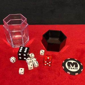 May include: A collection of magic-themed items on a red surface. Includes a black and white die, several white dice, a red die, a black octagonal container, a clear plastic container, and a casino chip with the text "Magic".