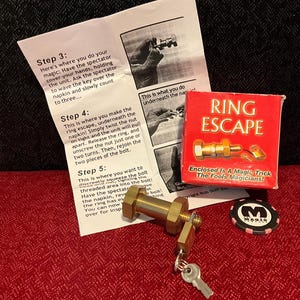 May include: A brass bolt and nut magic trick set including a red box labeled RING ESCAPE, an instruction sheet with steps 3, 4, and 5, a small padlock with a silver key, and a black and pink circular token with a white M logo in the center.