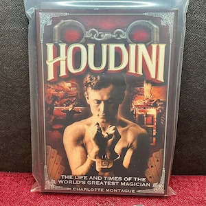 May include: A book titled Houdini: The Life and Times of the World's Greatest Magician by Charlotte Montague, featuring a shirtless man in handcuffs on the cover, enclosed in a clear plastic bag, accompanied by a small round black and pink token.