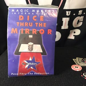May include: A Magic Makers International Dice Thru The Mirror magic trick set in a purple plastic bag. The packaging features the text Dice Thru The Mirror, an illustration of a die on a mirror, the Magic Makers logo, and the text Pass-Thru The Reflection.