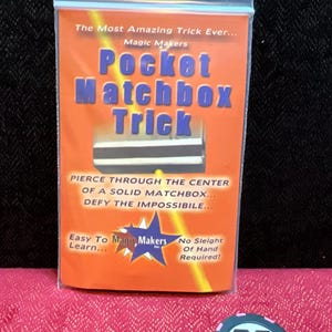 May include: A clear plastic bag containing a Pocket Matchbox Trick card with orange background and blue text. The card reads: The Most Amazing Trick Ever, Magic Makers, Pocket Matchbox Trick, Pierce through the center of a solid matchbox, defy the impossible, Easy To Learn, No Sleight Of Hand Required. A round black and pink chip with a white M logo is nearby.