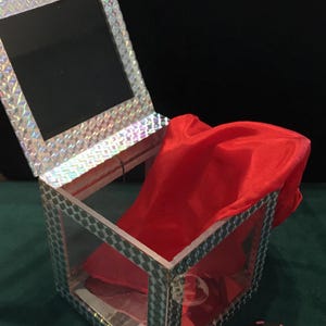 May include: A square box with clear sides and a silver holographic diamond-patterned frame. The box features an attached upright lid with a matching holographic border and a reflective interior. A bright red fabric cloth is draped inside the box.
