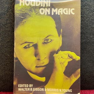 May include: A paperback book titled Houdini on Magic, edited by Walter B. Gibson and Morris N. Young, featuring a yellow-toned portrait of Harry Houdini on the cover. The book is enclosed in a clear plastic bag next to a round black and pink token.
