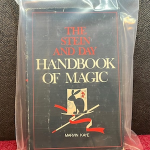 May include: A hardcover book titled The Stein and Day Handbook of Magic by Marvin Kaye, enclosed in a clear plastic bag. The black cover features red and white text, a graphic of a rabbit on a top hat, and a red ribbon. A small black poker chip is nearby.
