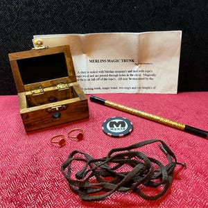 May include: A wooden magic trunk set including a small brown chest with a metal latch, a gold glitter magic wand with black tips, two gold rings with red circular insets, a black and pink magic token, a brown cord, and an instruction sheet.