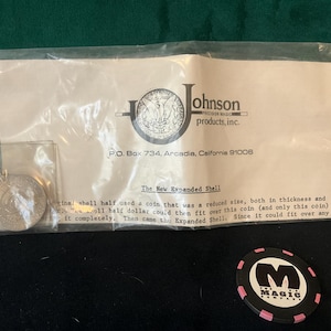 May include: A close-up of a white paper bag with the text "Johnson Products, Inc." and "P.O. Box 734, Arcadia, California 91006". The bag contains a silver coin and a black and pink poker chip with the word "MAGIC" printed on it.