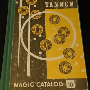 May include: A green hardcover book titled Louis Tannen Magic Catalog 10. The cover features a graphic design with a black and yellow background, white circular lines, and multiple illustrations of round coins with square center holes.