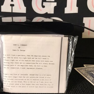 May include: A magic trick instruction sheet titled Zero G. Currency by James B. George, enclosed in a clear plastic sleeve. The white paper features black typewritten text describing a levitation illusion. A black object sits atop the sleeve.