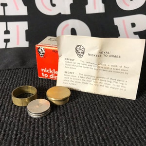 May include: A magic trick kit called 'Royal' Nickels to Dimes. The kit includes a brass ring, a brass cover, and a fake shell. The instructions on the card explain how to perform the trick.