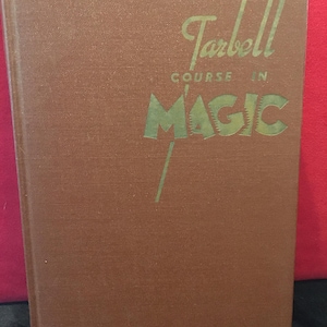 May include: A brown hardcover book titled Tarbell Course in Magic with gold lettering on the front cover. The text is arranged with the word Tarbell in script above the words COURSE IN and the large, bold, capitalized word MAGIC below it.