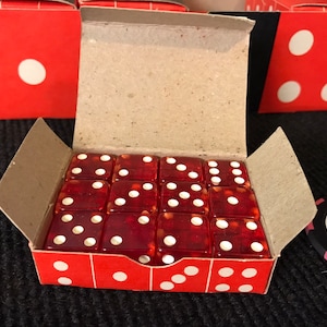 May include: A red cardboard box with a white dot on the front, open to reveal a set of 16 red dice with white dots. The dice are arranged in a 4x4 grid. The box is sitting on a black surface.