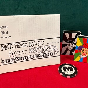 May include: A white envelope with the text "Matchbox Magic from... Bob Ostlin Clear Deception!"  Two matchbox-sized boxes with colorful designs, one with a black and white jester and the other with a colorful jester. A black and white chip with the letter "M" and the word "Magic" printed on it.