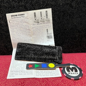 May include: A Dream Paddle magic trick set by John Kennedy Magic including an instruction sheet, a black velvet pouch, a black plastic paddle with a red star, green triangle, blue square, and yellow circle, and a round black and pink poker chip.