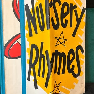 May include: A hardcover book titled Nursery Rhymes with a white cover, a yellow graphic element, and black star illustrations. The book features bold black text and a blue spine. Next to the book is a small black and pink circular token with a white M.