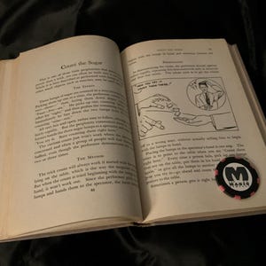 May include: An open book with black text on white pages titled Count the Sugar, featuring a black and white illustration of a magic trick. A round black and pink poker-style chip with the text The Magic Company is placed on the right page.