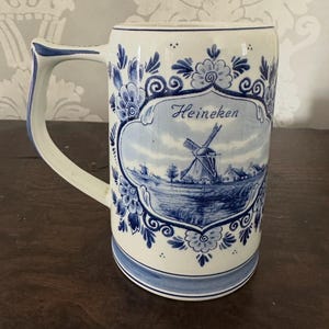 May include: A white ceramic beer stein with blue floral and windmill designs. The stein has a rectangular shape with a curved handle and the word "Heineken". The design includes a landscape scene with a windmill.