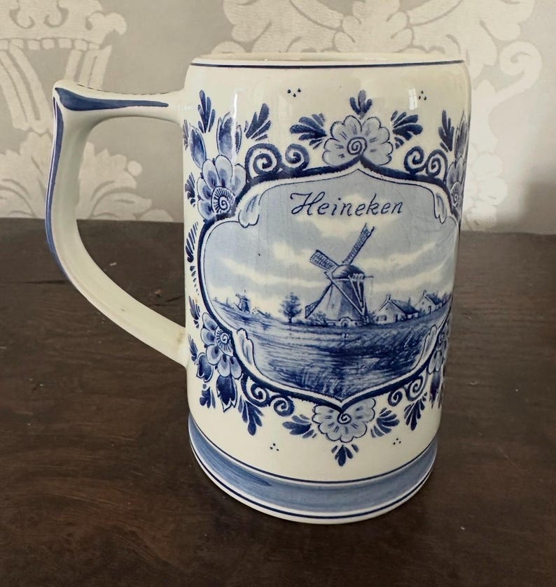 May include: A white ceramic beer stein with blue floral and windmill designs. The stein has a rectangular shape with a curved handle and the word "Heineken". The design includes a landscape scene with a windmill.