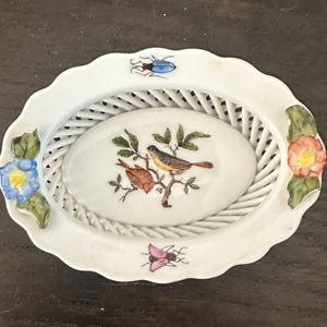 May include: White ceramic oval basket with a lattice design center. The center features a painted image of two birds on a branch. The rim is scalloped and decorated with blue and pink flowers and insects. A decorative piece.