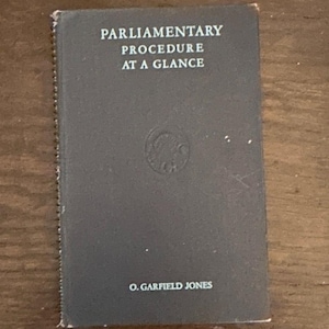 May include: A dark grey spiral-bound book titled Parliamentary Procedure at a Glance by O. Garfield Jones. The cover features white text and a circular embossed emblem in the centre. The book has a vertical wire binding along the left side.