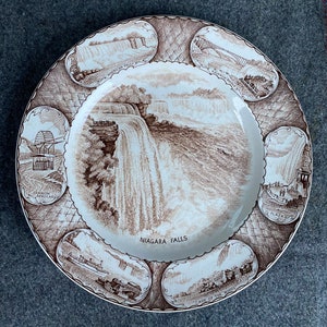 May include: A decorative plate featuring a sepia-toned illustration of Niagara Falls. The plate's center showcases the falls, surrounded by vignettes of landmarks like the Rainbow Bridge and the Maid of the Mist. The words "Niagara Falls" are printed on the plate.