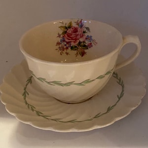 Può includere: Una tazza da tè e un piattino bianchi con un motivo floreale. La tazza ha un bordo di vite verde e una rosa rosa con foglie verdi e fiori blu al centro. Il piattino ha un bordo di vite verde e un bordo ondulato.