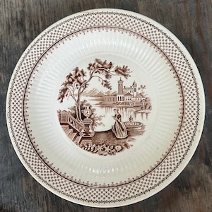Könnte beinhalten: Ein weißer Keramikteller mit braunem Rand und einem braunen Bild einer Landschaft mit einem Gebäude, einem Brunnen und einer Frau in einem Kleid. Der Teller hat einen gewellten Rand.