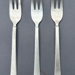May include: Three silver-colored forks with a simple design. The forks have a slightly textured finish and are arranged in a row.