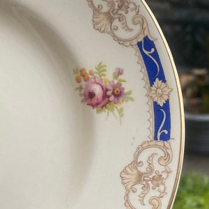 Pode incluir: Um prato de cerâmica branco com uma borda azul e dourada. A borda apresenta um desenho floral com flores rosa e amarelas. O prato tem uma borda dourada.