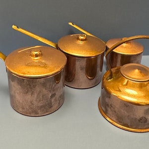 Puede incluir: Conjunto de cuatro cacerolas de cobre en miniatura con asas de latón. Las cacerolas tienen tapas y están en acabado pulido. Una olla es una tetera con asa.