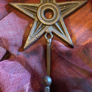 May include: A cast iron star-shaped hook with a ring at the top and a rounded hook at the bottom. The hook is painted in a dark brown color and has a rustic look.