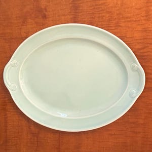 Puede incluir: Una fuente ovalada azul claro con asas decorativas. La fuente tiene una superficie lisa y un borde ligeramente elevado. Las asas presentan un diseño circular. La fuente está sobre una superficie de madera marrón.