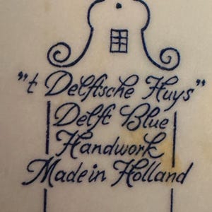 May include: A close-up of a white surface with a blue illustration of a house and text. The text reads "'t Delfsche Huys", "Delft Blue Handwork Made in Holland". The illustration is in a traditional Dutch style.