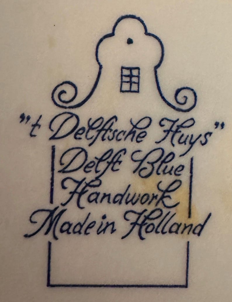 May include: A close-up of a white surface with a blue illustration of a house and text. The text reads "'t Delfsche Huys", "Delft Blue Handwork Made in Holland". The illustration is in a traditional Dutch style.