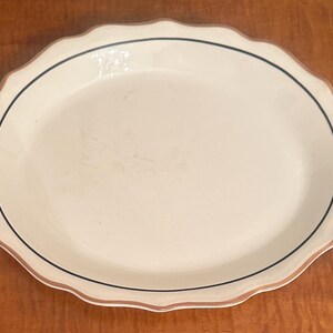 Puede incluir: Una fuente ovalada con borde festoneado. El plato de cerámica blanquecina tiene una fina línea negra y un borde marrón alrededor del borde. La fuente está vacía y descansa sobre una superficie de madera.