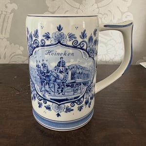 May include: A white ceramic beer mug with blue floral patterns and a handle. The front features a blue and white illustration with the word "Heineken". The mug has a wide base and a curved handle.
