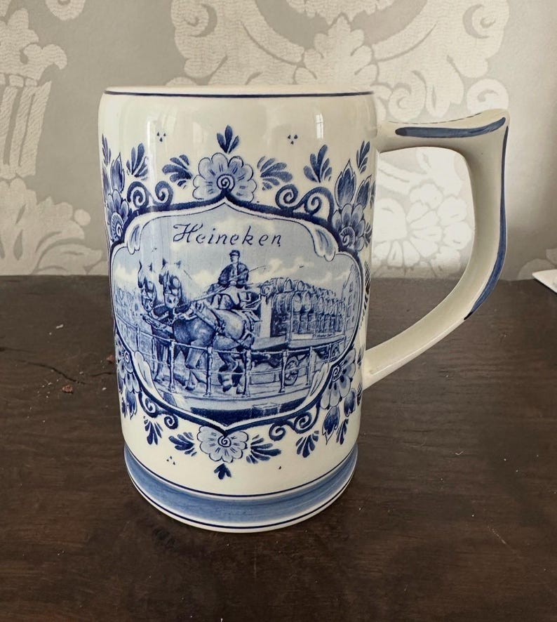 May include: A white ceramic beer mug with blue floral patterns and a handle. The front features a blue and white illustration with the word "Heineken". The mug has a wide base and a curved handle.
