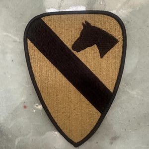 May include: A tan and black embroidered patch with a shield shape. The patch features a black diagonal stripe and a black horse head silhouette in the upper right corner. The patch has a black border.