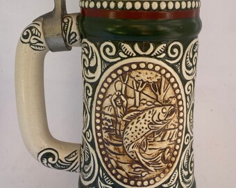 May include: A ceramic beer stein with a green and white design. The stein features a scene of a fisherman catching a fish. The text "The Strike...Rainbow Trout" is printed on the bottom of the stein.