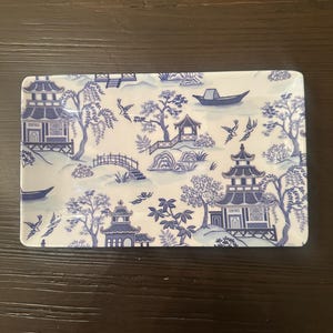 Puede incluir: Bandeja rectangular de cerámica con un diseño azul y blanco de estilo chinoiserie. El patrón presenta pagodas, árboles, puentes, barcos y pájaros. La bandeja es rectangular y tiene esquinas redondeadas.
